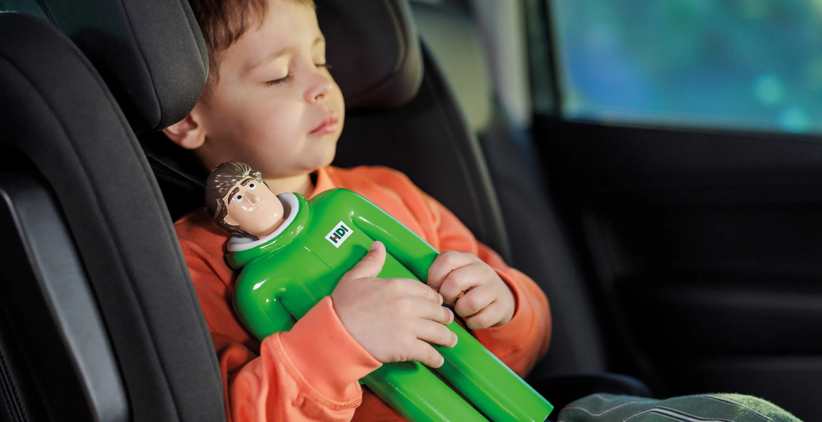 Kind sitzt in einem Kindersitz im Auto und hält eine grüne HDI-Figur im Arm.