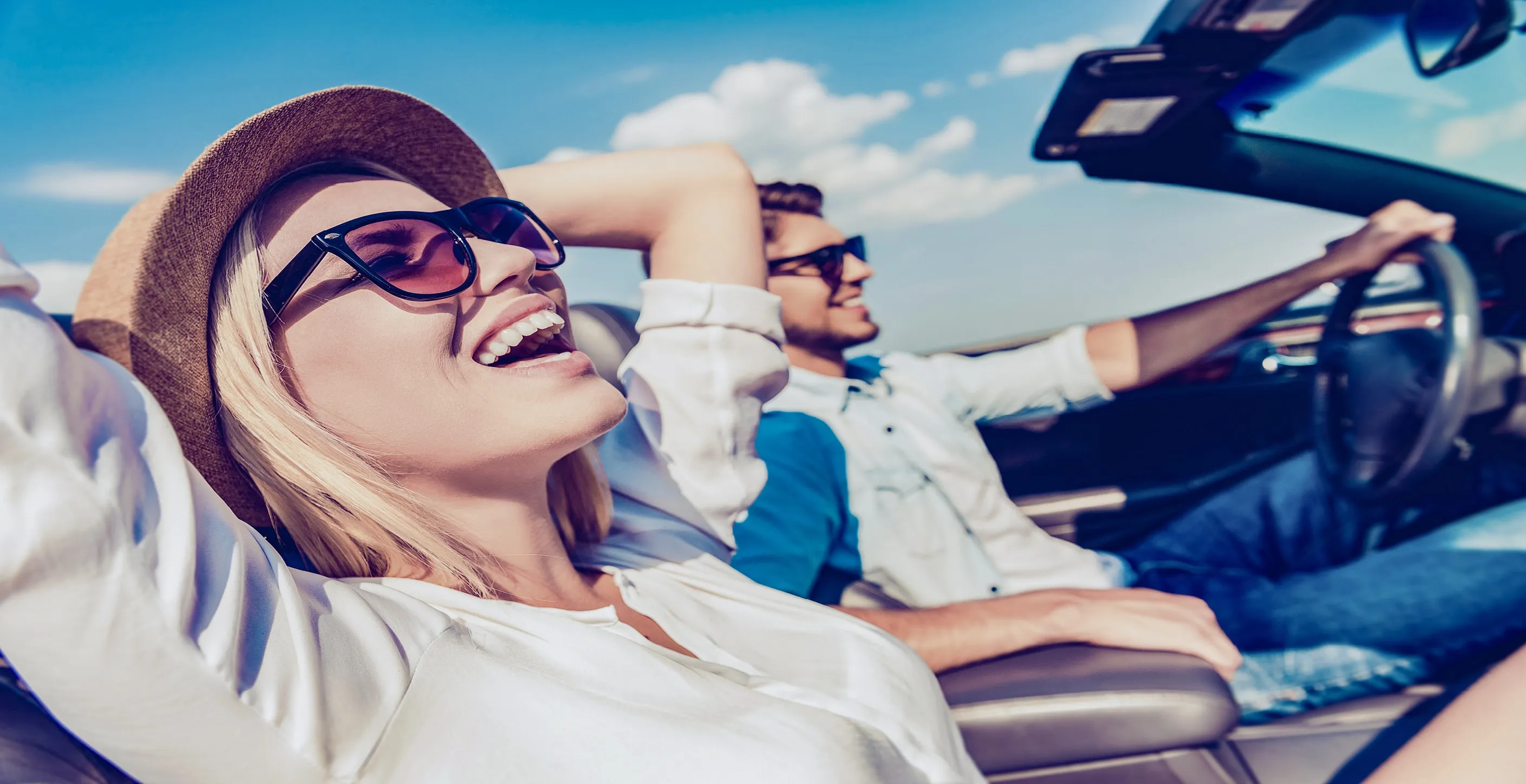 Zwei Personen sitzen in einem Cabrio und fahren auf einer Straße bei sonnigem Wetter